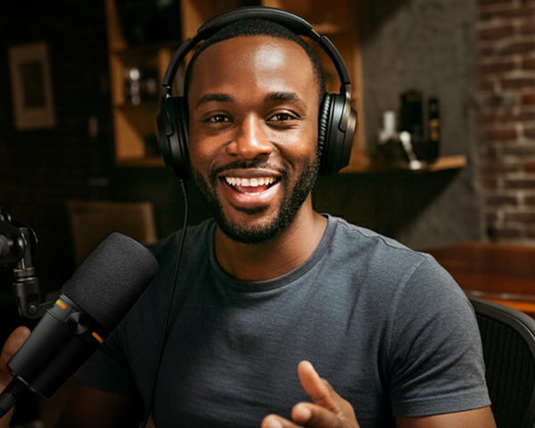 Ein lächelnder Mann mit Kopfhörern sitzt vor einem Mikrofon in einem gemütlichen Studio und spricht, während er einen Podcast aufnimmt.